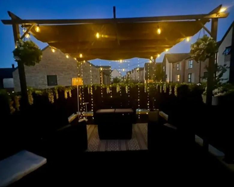 Night view of a decorated balcony with a pergola, lights, and seating.