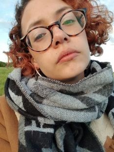 Close-up portrait of a young woman with curly red hair, wearing glasses and a scarf.