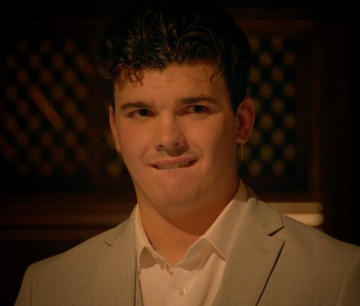 Harry Clark, in a light suit and white shirt, smirks while biting his lip.