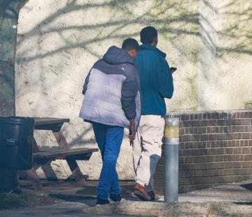 Two men walking near a brick wall.