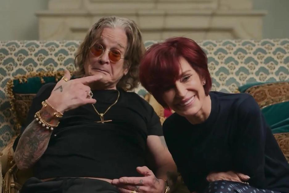 Sharon and Ozzy Osbourne sitting on a patterned couch, with Ozzy pointing at himself and Sharon smiling.
