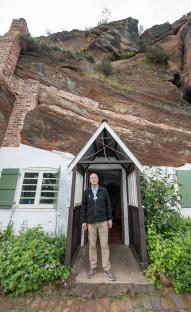 A man stands in the doorway of a house carved into a sandstone cliff.