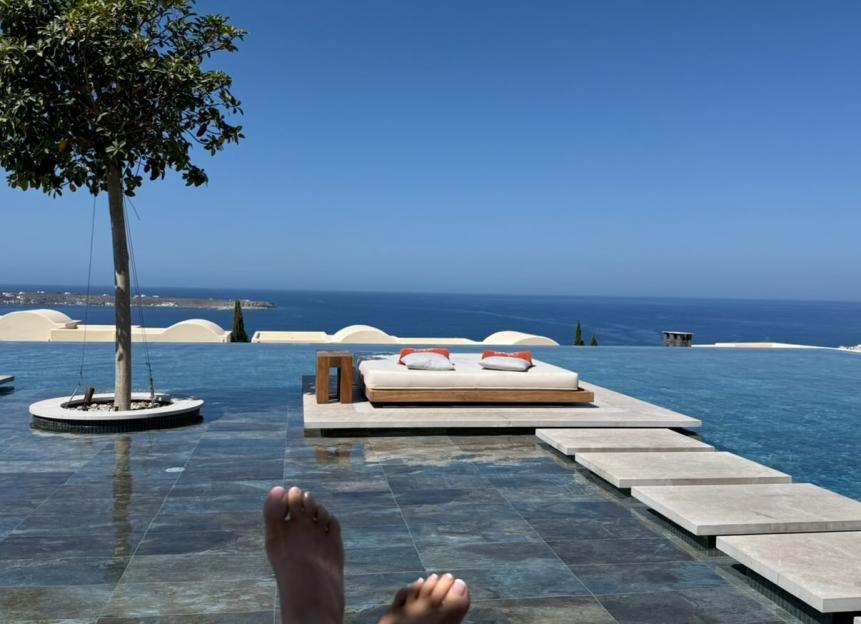 Bare feet with pink toenail polish relaxing by an infinity pool in Santorini.