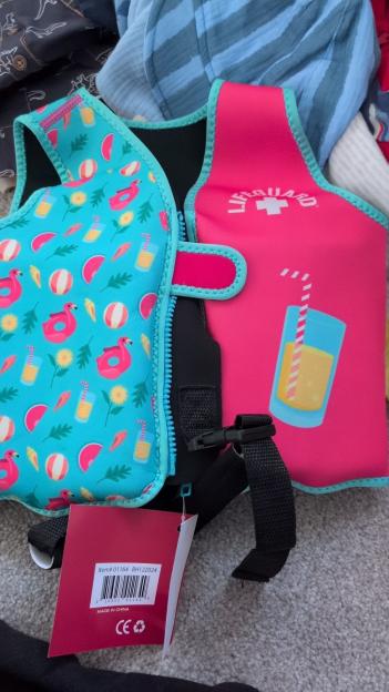 Close-up of two children's swim vests, one blue with flamingo and beach ball patterns and the other pink with "LIFEGUARD" and a drink illustration, both with an attached tag.