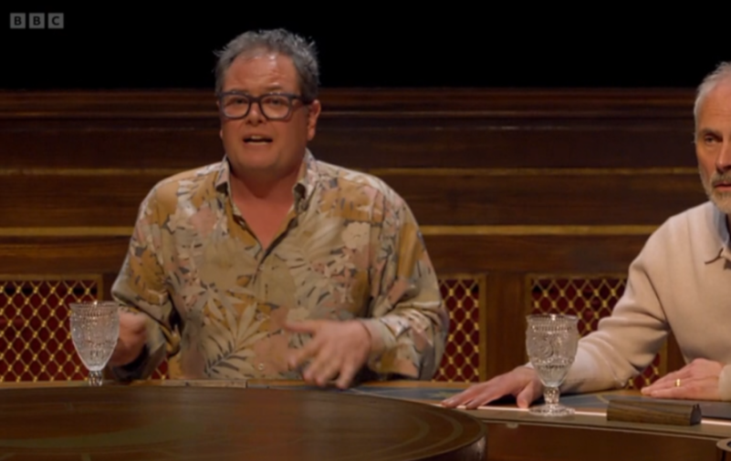 Alan Davies speaking at a table with an unknown man beside him.