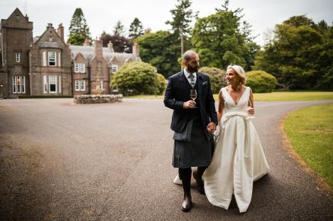 Newlyweds holding hands and wine glasses, walking away from a large stone house.