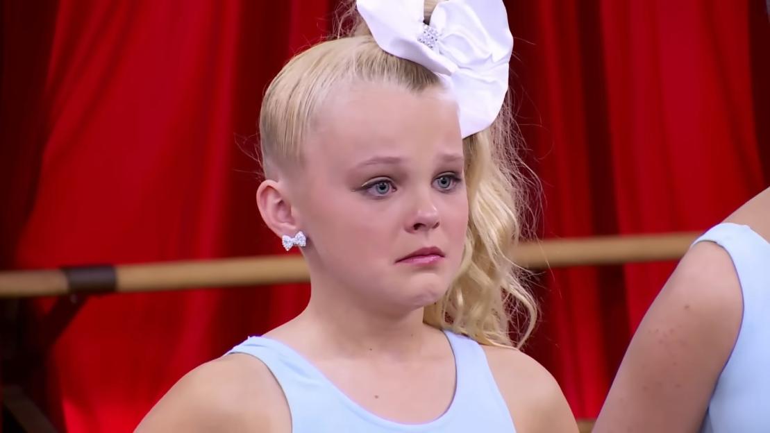 JoJo Siwa crying, wearing a large white bow, with blonde hair in a high ponytail.