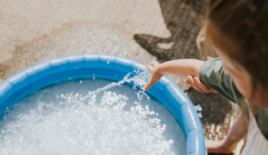 A girl's hand touches the water spraying from a garden hose into a blue inflatable pool.