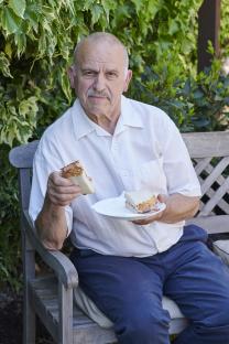 Older man sitting on a bench outdoors, eating a bacon sandwich.