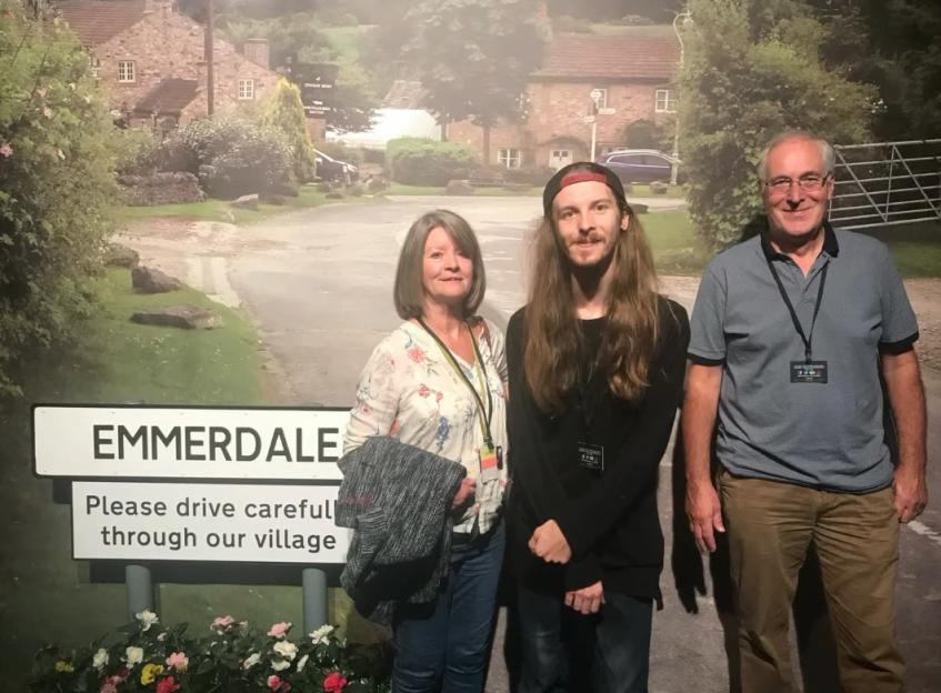 Three people posing in front of an Emmerdale village backdrop.