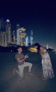 A man proposing to a woman on a beach at night with city skyline in the background.