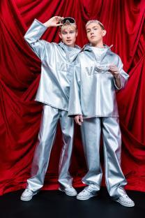 Two people in matching silver outfits and jeweled sneakers pose in front of a red curtain.
