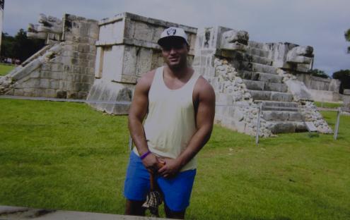Man in tank top and shorts standing in front of Mayan ruins.