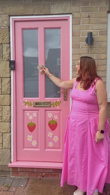 Woman in pink dress standing by a pink door decorated with strawberries and flowers.