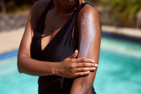 Close-up of a Black woman applying sunscreen to her arm by a pool.