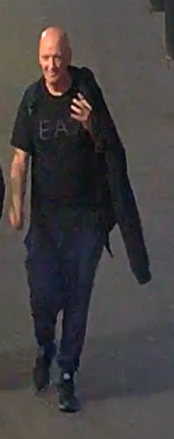 Surveillance image of a bald man walking, wearing a black t-shirt and dark pants.