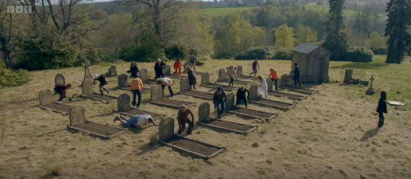 People dressed in various outfits emerge from graves in a graveyard.