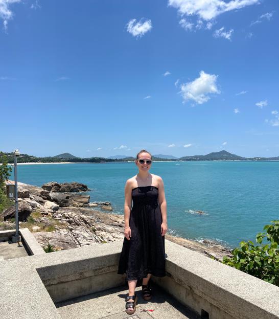 A woman in a black dress standing on a stone balcony overlooking a blue ocean with mountains in the background.