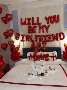 Romantic bedroom decorated with red balloons spelling "Will you be my girlfriend Abi Love" and towel swans.