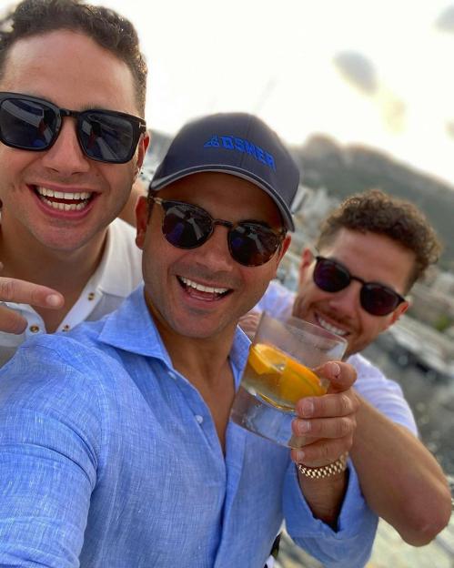 Three men in sunglasses smile at the camera, one holding a drink.