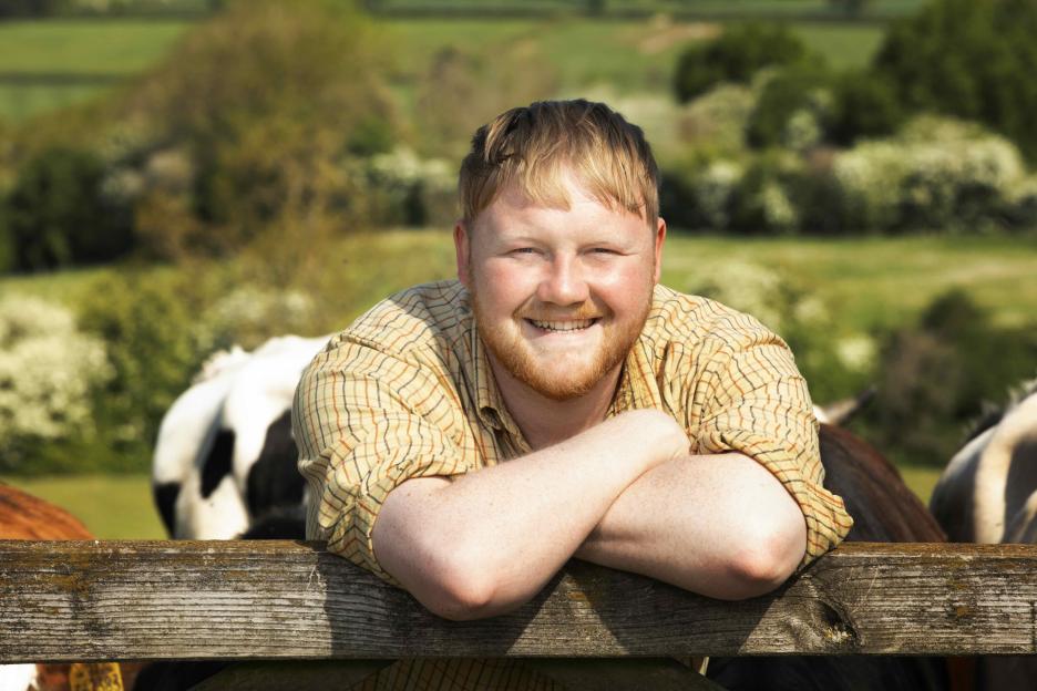 Kaleb Cooper in an olive green long-sleeved polo shirt with the initials "KC" embroidered, blue jeans, leaning on a fence post with a field and cloudy sky in the background.