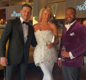 Three people at an event; two men in suits and a woman in a white dress.