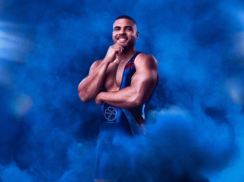 Portrait of Zack George, a muscular man in a wrestling singlet, posing against a blue smoky background.