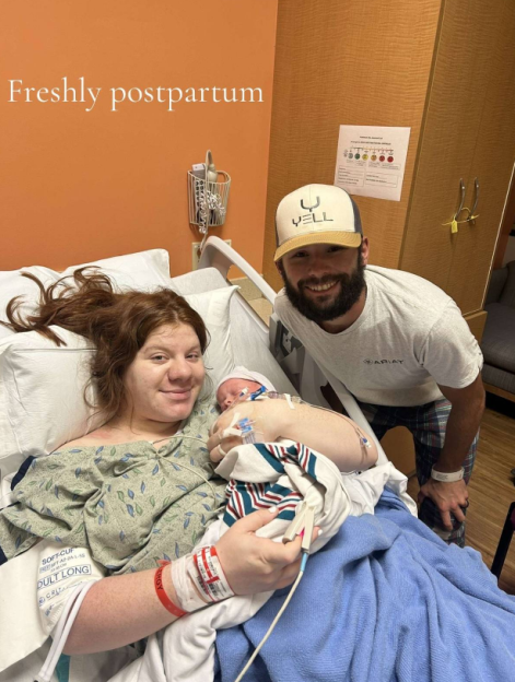 A woman holding a newborn baby in a hospital bed next to a smiling man in a baseball cap.