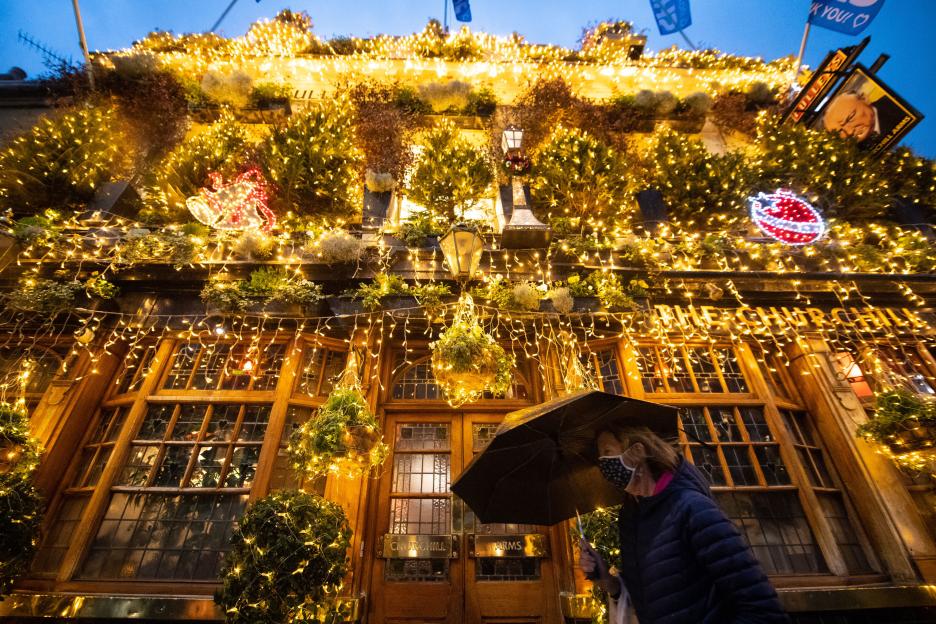Woman in a face mask walking past the closed Churchill Arms pub decorated with Christmas lights.