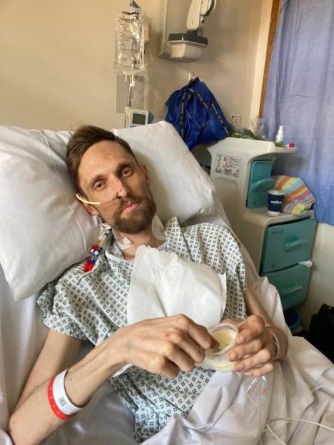 Ryan Long in a hospital bed with a nasal cannula, eating from a small container.