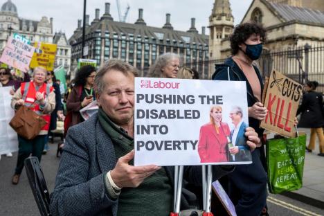 Protestor in wheelchair holding sign reading "Pushing the disabled into poverty" at a demonstration outside Parliament.