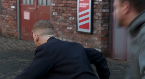 Screenshot of a man running away from another man in an alleyway.