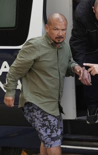 A handcuffed man in a green shirt and patterned shorts is escorted into a police vehicle.