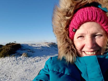 Close-up of a smiling woman in a teal coat and pink hat, outdoors in a snowy landscape.