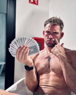 A shirtless man holds a fanned deck of cards and looks unhappy.