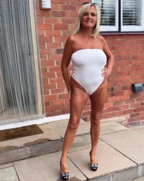 A woman in her 60s wearing a white swimsuit and heels, posing outdoors.