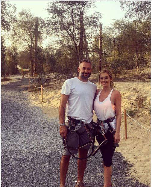 A man and woman wearing harnesses stand on a gravel path next to a sandy area with trees in the background.