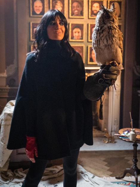 Presenter Claudia Winkleman with an owl perched on her arm, in a room with framed portraits on the wall.