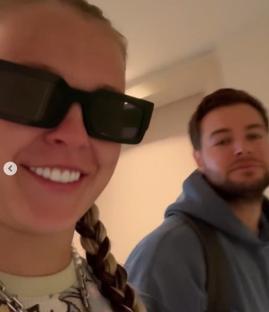 Close-up of a smiling woman wearing sunglasses and a man behind her.