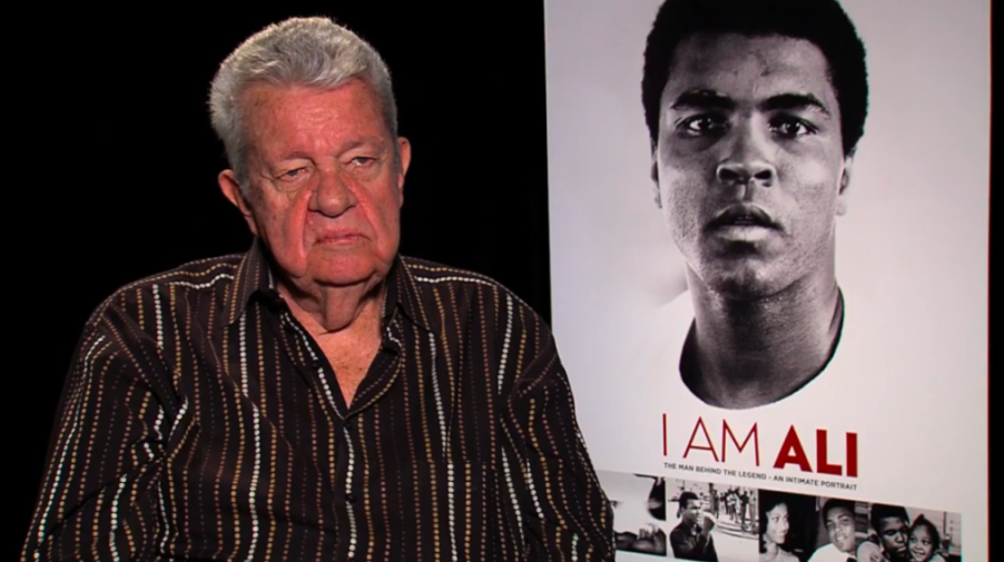 A man in a striped shirt sits next to a poster for the documentary "I Am Ali."