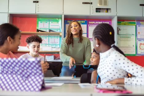 A Black female teacher helps diverse students using tablets in a classroom.