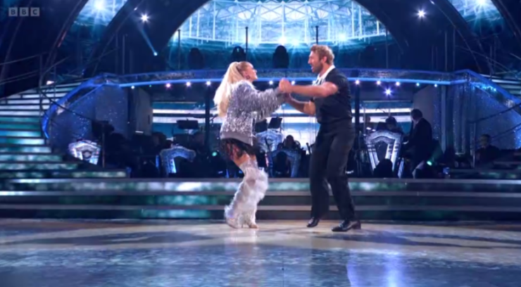 A woman in a sparkly silver top and white feathery leg warmers dances with a man in a black shirt and pants on a brightly lit stage.