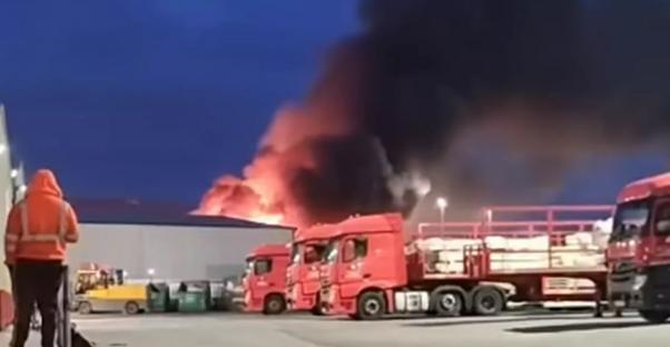 Large fire engulfing a building with several red semi-trucks parked nearby.
