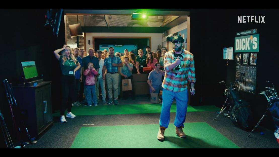 Adam Sandler in a golf simulator, surrounded by onlookers.