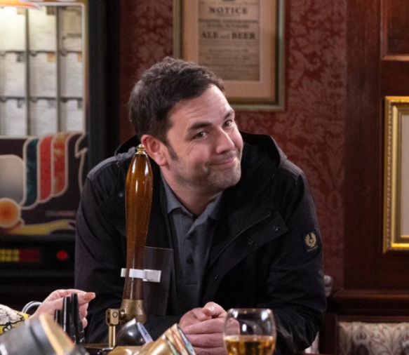 A man with dark hair in a black jacket and gray shirt smiling, with beer taps and a glass of beer in front of him.