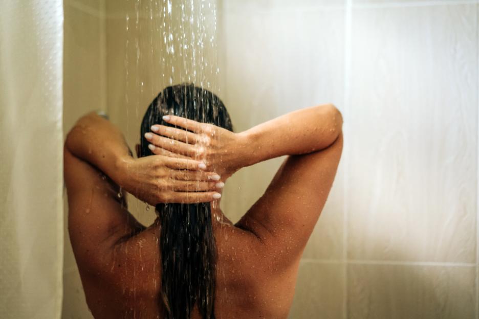 A woman showering in a hotel room with her hands touching the back of her head, seen from behind.