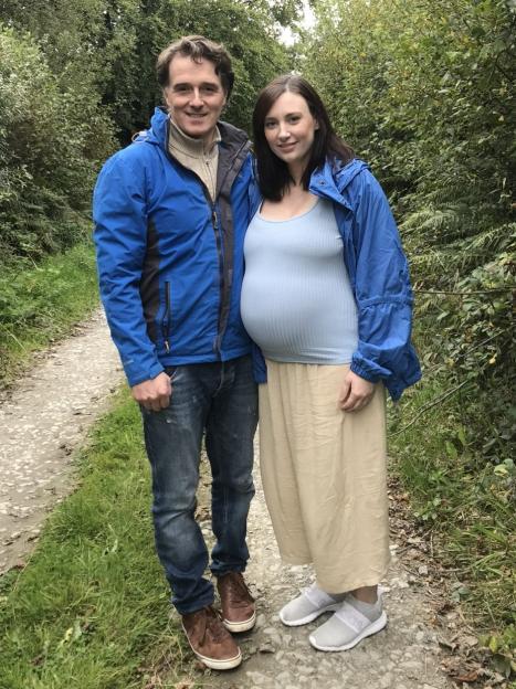 A man and a pregnant woman stand on a path surrounded by green foliage.