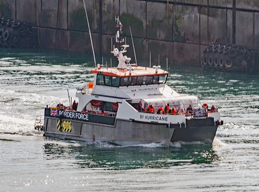 A Border Force vessel, "BF Hurricane," with rescued migrants in orange life jackets, moving through water at Dover Docks.