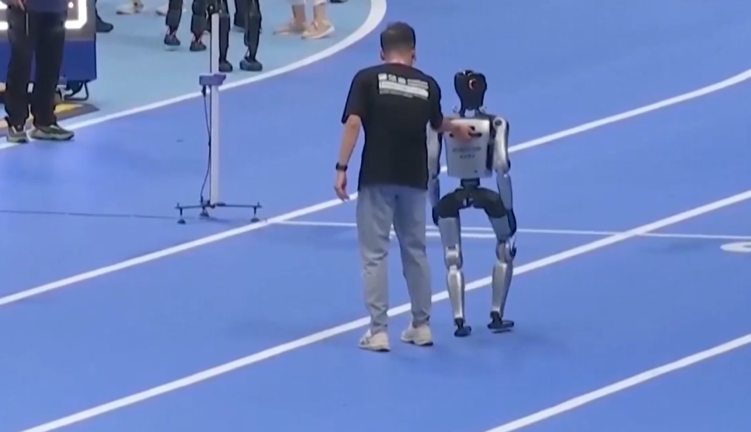 A person assists a bipedal robot walking on a blue track.