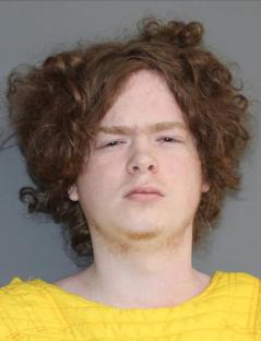 Mugshot of a young man with curly brown hair wearing a yellow shirt.